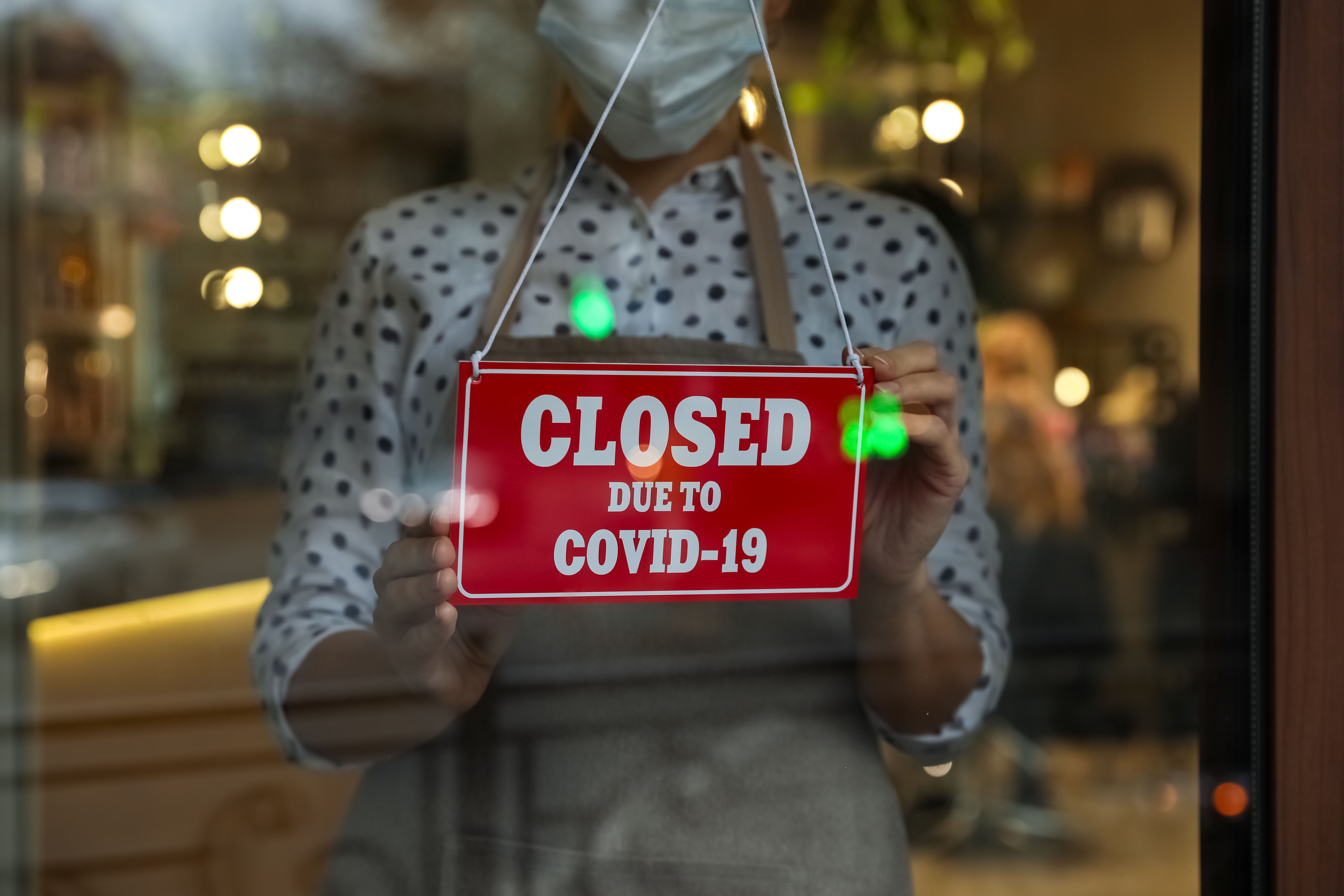 Woman in mask putting red sign with words Closed Due To Covid-19 hanging onto glass door, closeup